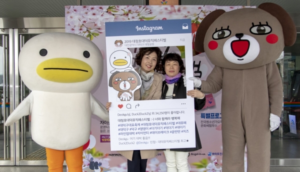 대덕구청 현관 앞에 설치된 대청호대덕뮤직페스티벌 포토존 앞에서 포즈를 취하고 있는 주민과 대덕구 캐릭터 덕구(오른쪽)과 더기더기(왼쪽)]